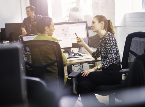 Two women talking at a desk in front of a computer screen