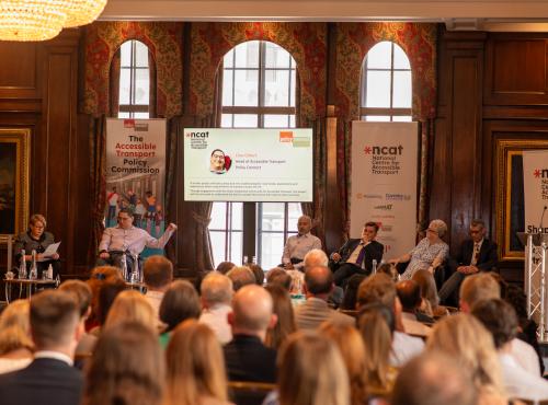 Launch of the Accessible Transport Policy Commission. Photo taken behind the audience - looking at the stage where speakers and panellists are seated. Two banners on either side of a screen with a powerpoint presentation