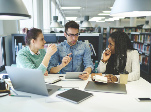 Three people discussing work at a desk with technology around them including a two laptops and a phone